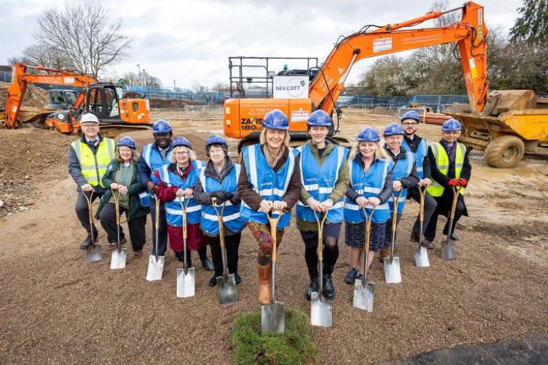 Eleven people wearing hard hats stood on soil with their feet on spades