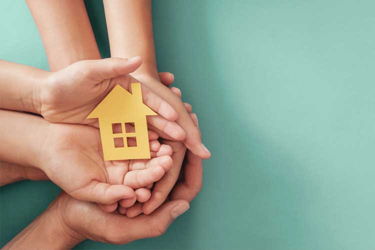 Several hands of different sizes are stacked together, holding a small yellow paper cutout of a house with four windows. The background is a solid teal color.