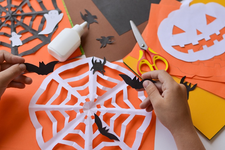Hands making Halloween crafts with paper bat, spider web, and themed cutouts on a table.