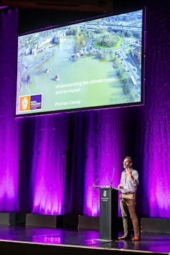 Professor Ian Candy stands on a stage lit in purple lights in front of a large screen. The screen shows a photo of a flood and reads "Understanding the climate crisis and its impact".