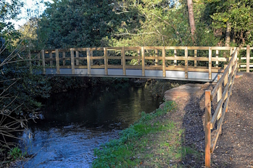 A bridge made from pale wood with a sturdy metal base going across a river. The bridge has safety rails to either side, and a fence has been placed beside the river bank.