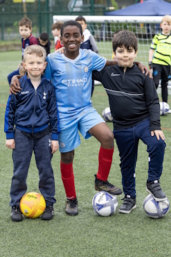 A group of three boys stand on a football pitch, with footballs at their feet.