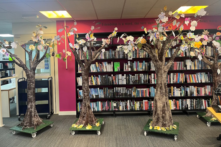Four handmade trees in front of a row of bookshelves in Bracknell Forest Library. Each tree is covered in leaves which have been individually decorated.