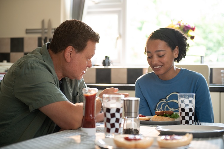 Two people sit at a kitchen table eating and talking, with food, drinks, and condiments in front of them.