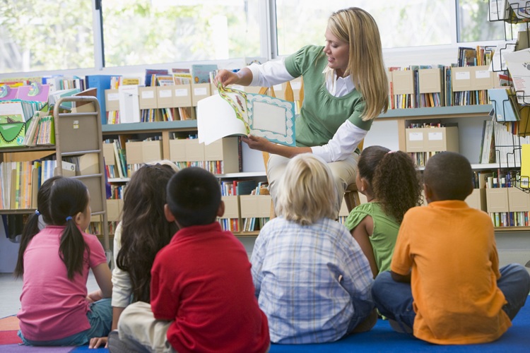 A person reads a picture book to a group of children sitting on the floor in a library