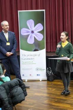 A primary school aged girl stands in front of an audience with a microphone. Behind her is a banner which reads "the principle of geometry".