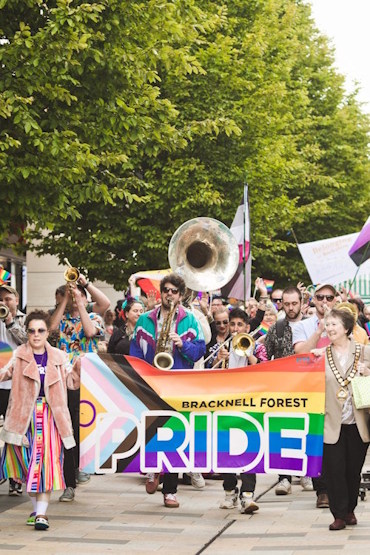 Bracknell Forest Pride's parade. Two women hold up a banner of the Progress Pride flag. Behind them is a crowd of people led by a brass band.