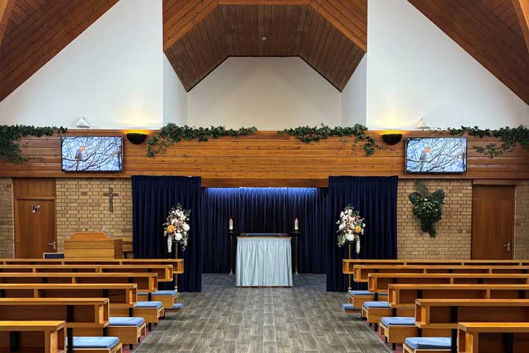 Chapel interior with wooden ceiling, rows of benches, and a central altar framed by curtains and floral arrangements