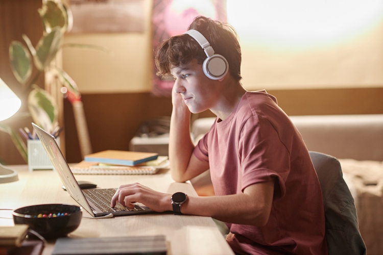 A young teenager sits at a desk working at a laptop with headphones on.