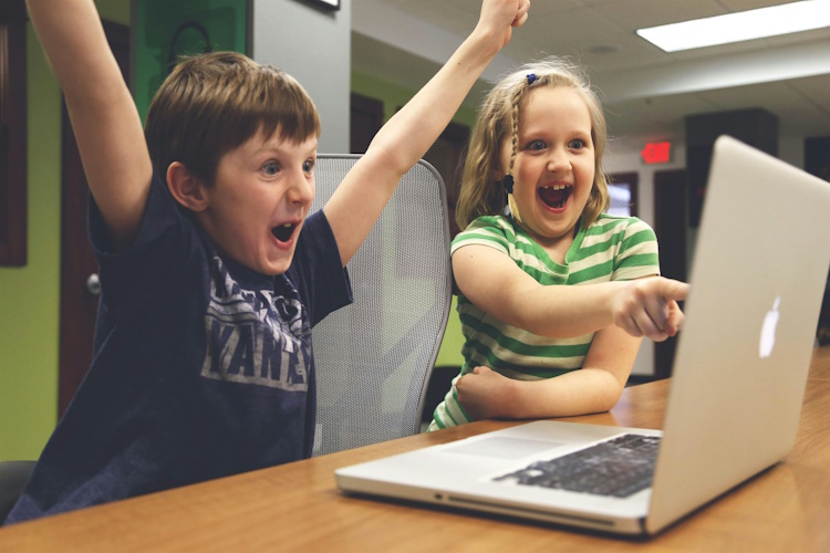 Two children sit in front of a laptop with excited expressions. One has their arms in the air in celebration while the other points at the laptop screen.
