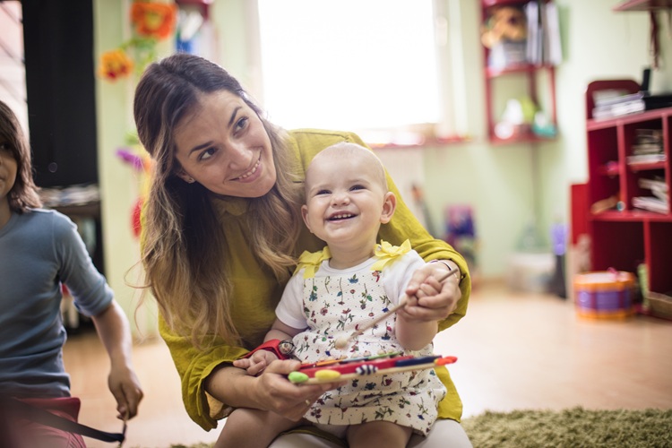 A woman is sitting on the floor with a baby on her lap. She is helping the baby to play a toy tambourine. Both the woman and baby are smiling.
