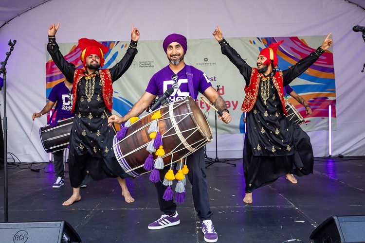 A sikh man in purple turban playing a dhol drum. Two male dancers in traditional dress dance with their arms in their air either side of him.