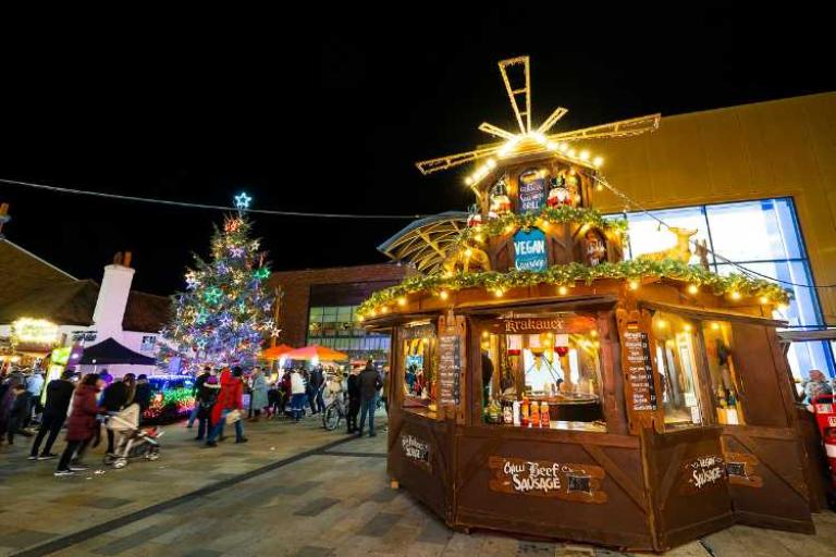 A christmas market stall with lights at night with a christmas tree in the background.,