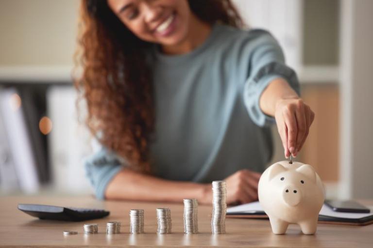 Woman in a grey top sitting behind a table, smiling as she puts coins in a pink piggy bank. Coins and a calculator are on the table next to the piggy bank 