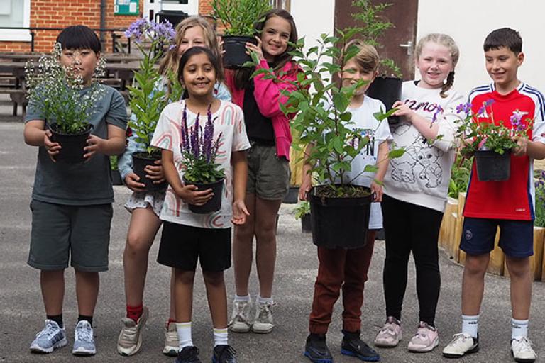 Seven primary school age children in outdoor clothes standing in a playground and holding a variety of small potted plants
