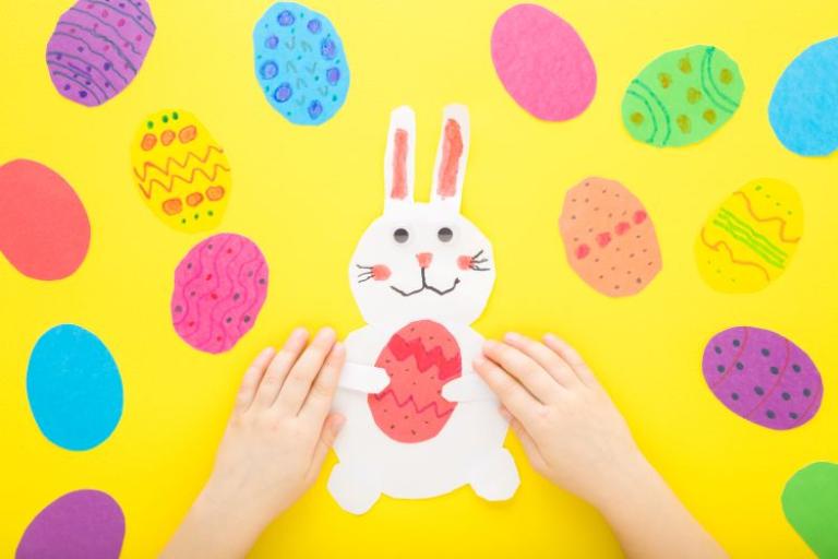 Child's hands on a paper Easter bunny holding an Easter egg, surrounded by colourful paper eggs