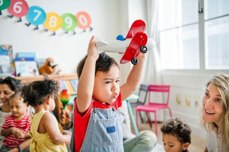 Toddler holding up red air plane in a busy playgroup