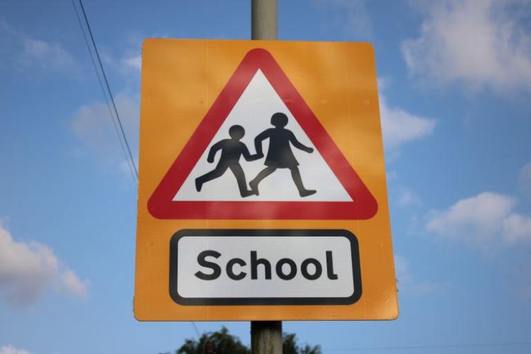 A red triangle road sign for school with two children in the middle, set against a bright blue sky with clouds. 