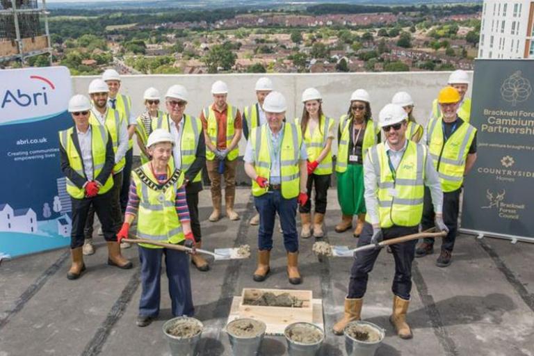 Group of people in high-vis jackets and hard hats on top of the roof of a tall building. They are standing in front of several buckets of cement and two people are holding shovels. 