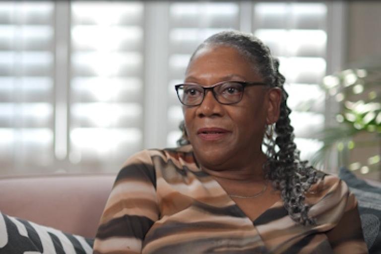 A woman sits on a sofa while giving an interview to camera.