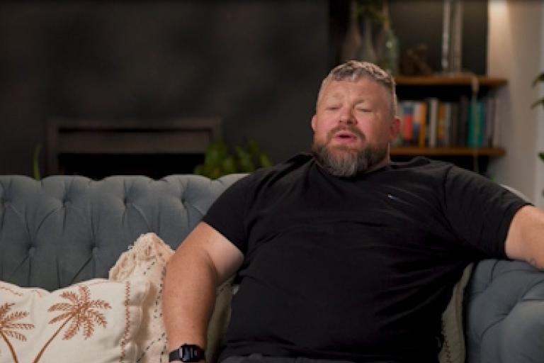 A man sits on a grey sofa giving an interview to camera.