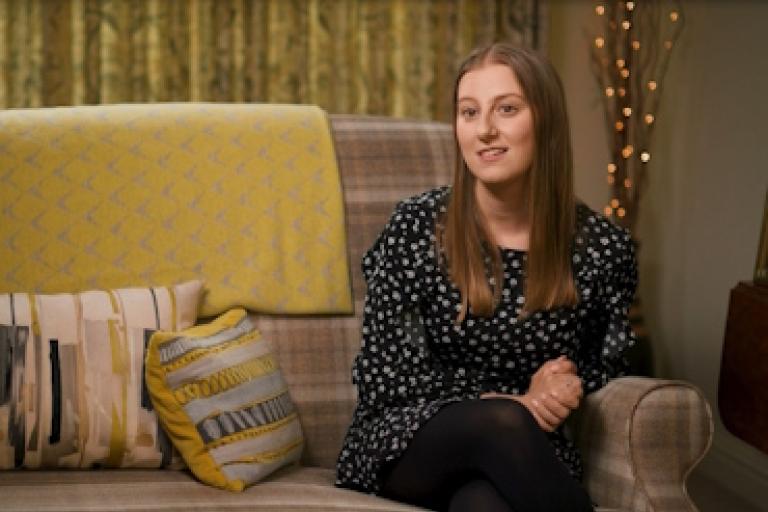 A woman sits on a sofa in a warm, comfortable looking room. She is giving an interview to camera.