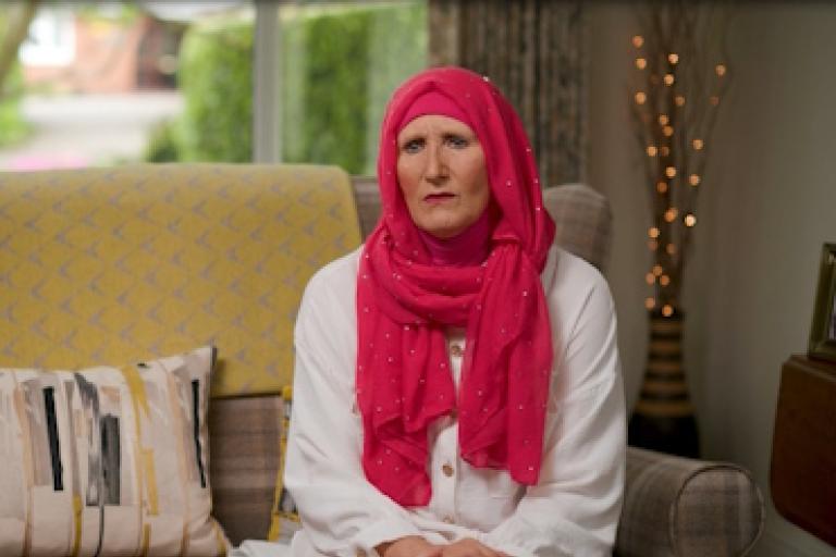A woman in a bright pink hijab sits on a sofa giving an interview to camera.