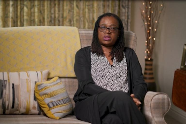 A woman sits on a beige sofa giving an interview to camera.