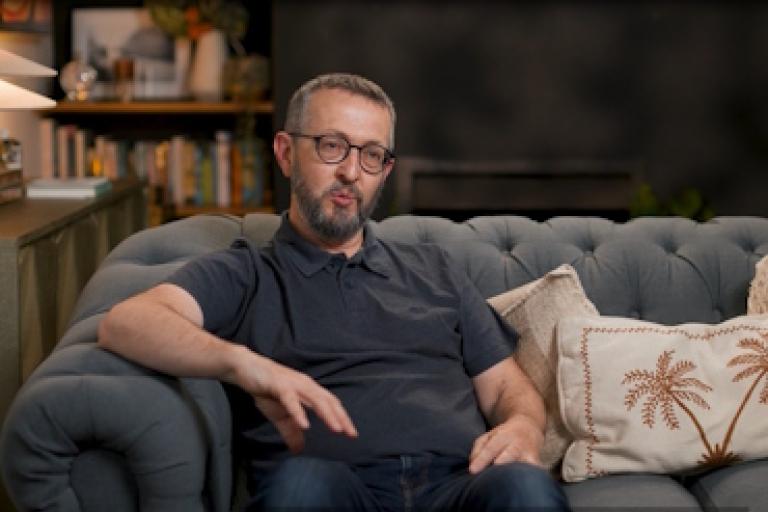 A man in a grey shirt sits on a sofa giving an interview to camera.