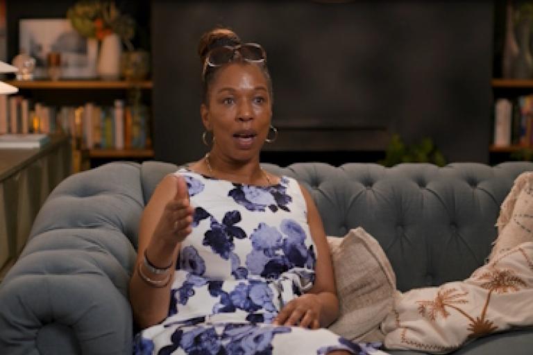 A woman in a floral dress sits on a sofa and gives an interview to camera.