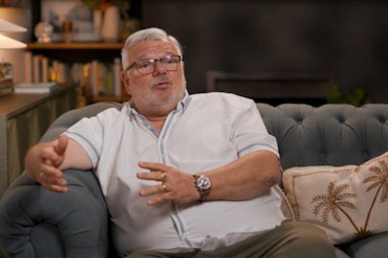 A man in a white shirt sits on a sofa giving an interview to camera.