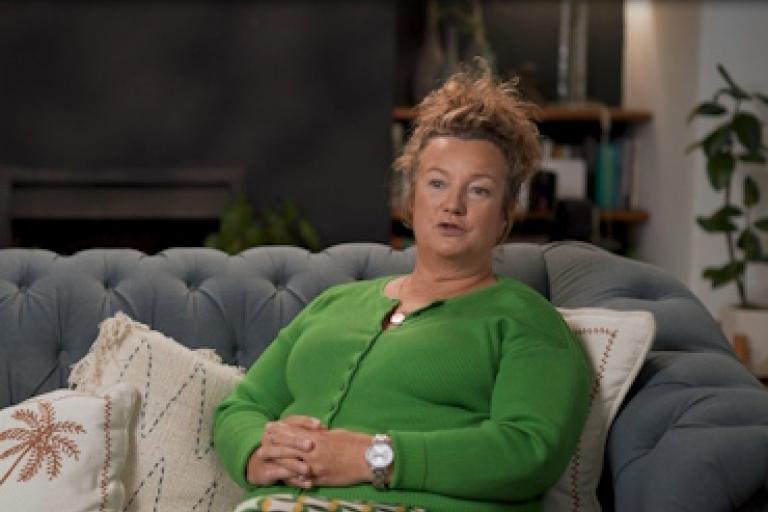 A woman in a green cardigan sits on a sofa and gives an interview to camera.