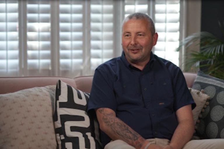 A man sits on a sofa smiling as he gives an interview to camera.