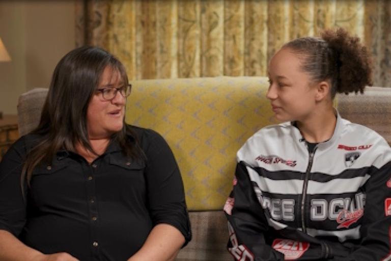 Two women sit on a sofa looking at each other. The woman on the left is older, and is speaking. The woman on the right is younger and is listening to her speak.