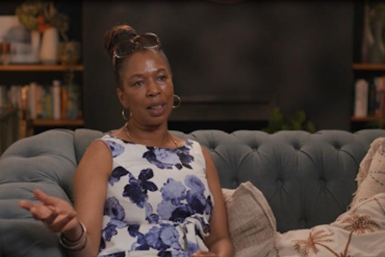 A woman in a floral dress sits on a sofa giving an interview to camera.