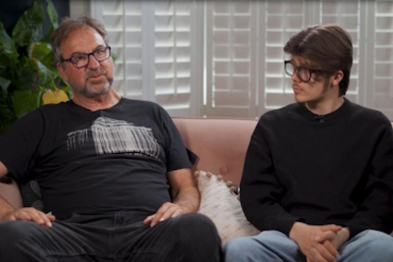 Two men sit on a sofa giving an interview. The man on the left is older and is speaking. The man on the right is younger and listens to him speaking.