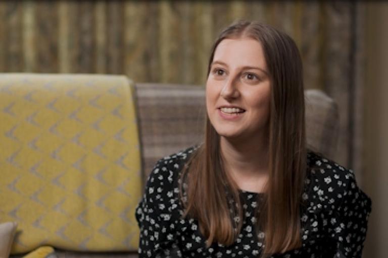 A smiling woman sits on a sofa giving an interview to camera.