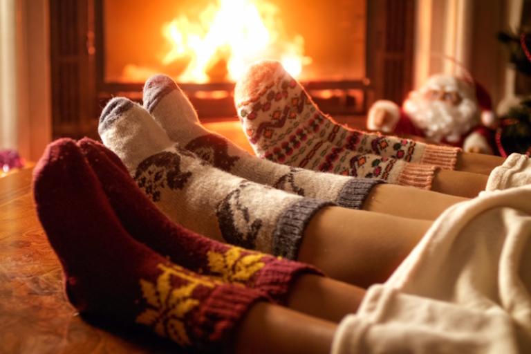 A close up on the feet of three people wearing Christmas themed socks with their feet up in front of a festive fireplace.