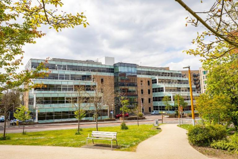 Exterior photograph of five storey office building with trees, grass and a bench in front of it. 