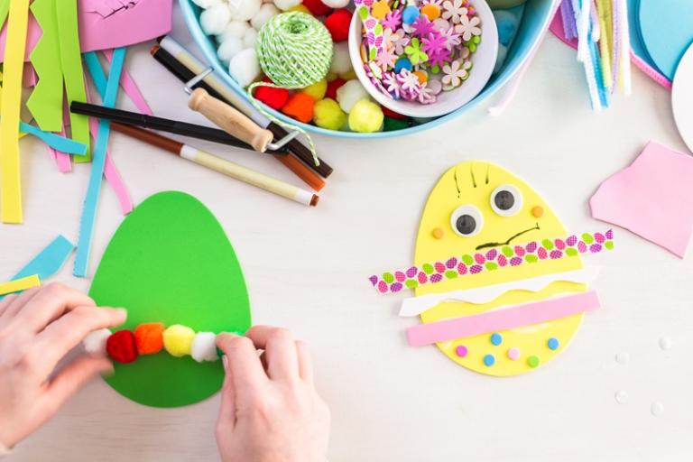 Hands decorating a green foam Easter egg with pom‑poms beside a finished yellow egg and assorted craft supplies on a table