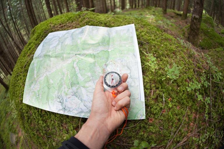 Hand holding a compass over a topographic map on a mossy rock in a forest