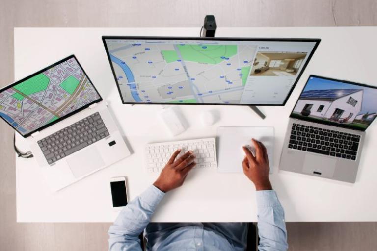 A man in a blue shirt at a computer that has maps on the screen. A laptop is either side of the main screen and have property images on them. 