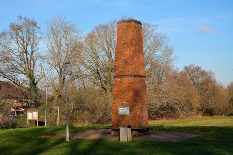 A replica brickworks chimney. This is a large, red brick chimney in the middle of a park.