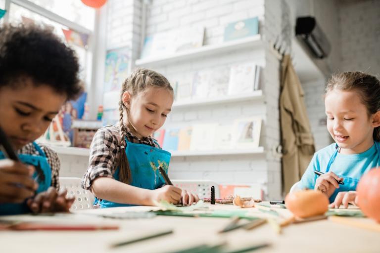 Children sit around a table in a library wearing aprons, doing crafts.