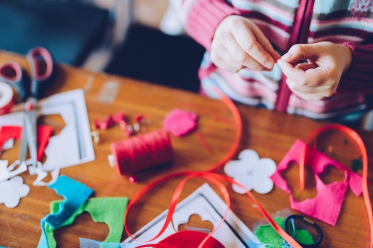 A child sits at a table covered in craft material like paper, beads and thread.