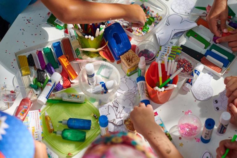 A table shot from above covered in various craft materials, including paints, glitter, stickers, pens and glue.
