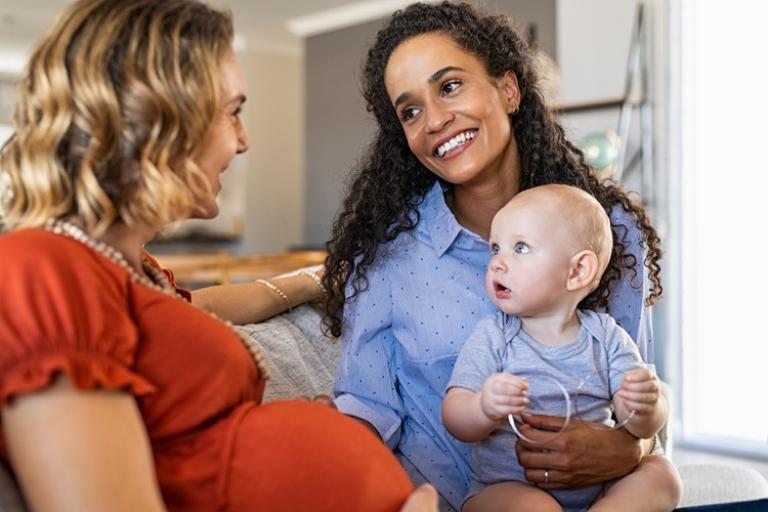 Two women are sat on a sofa talking. One is pregnant and the other sits with a baby on her lap facing her. The women are smilling at each other.