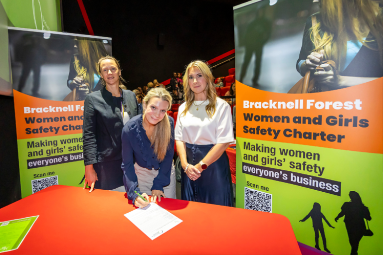 3 women in with banners beside them. One is signing a piece of paper on a table with an orange tablecover.