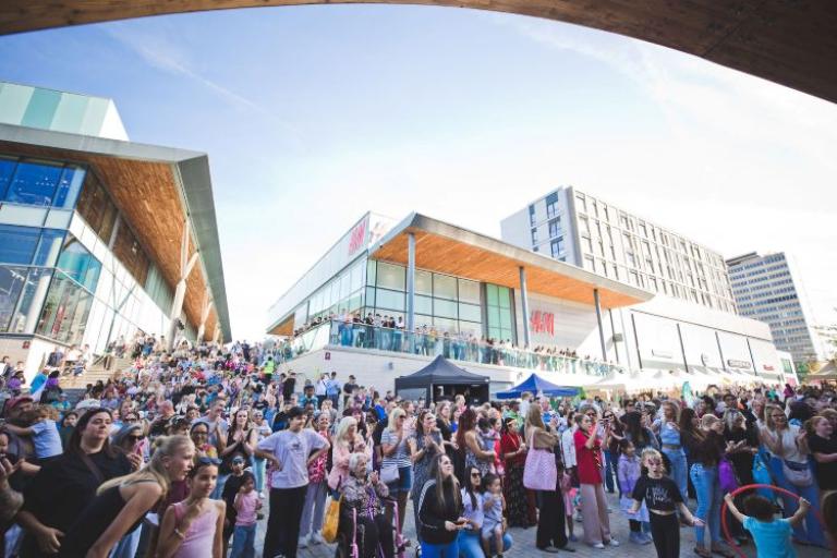 a crowd of people watching stage with a shopping centre behind them