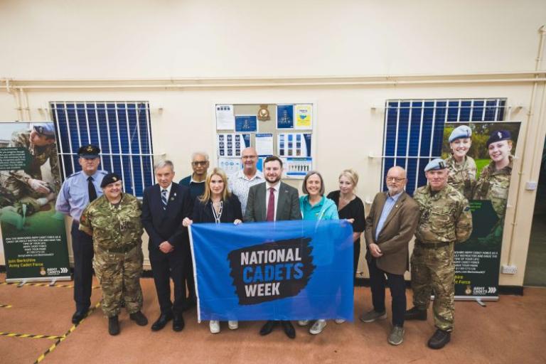 A group of people in a hall with some dressed in army and air force uniform. They are holding a big banner that says national cadets week